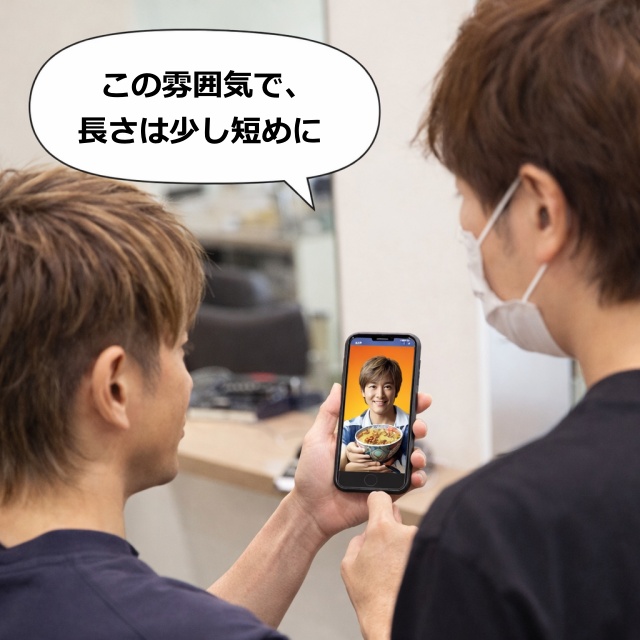 美容師にスマホ画面を見せながら「この雰囲気で、長さは少し短めに」とオーダーしている様子。
